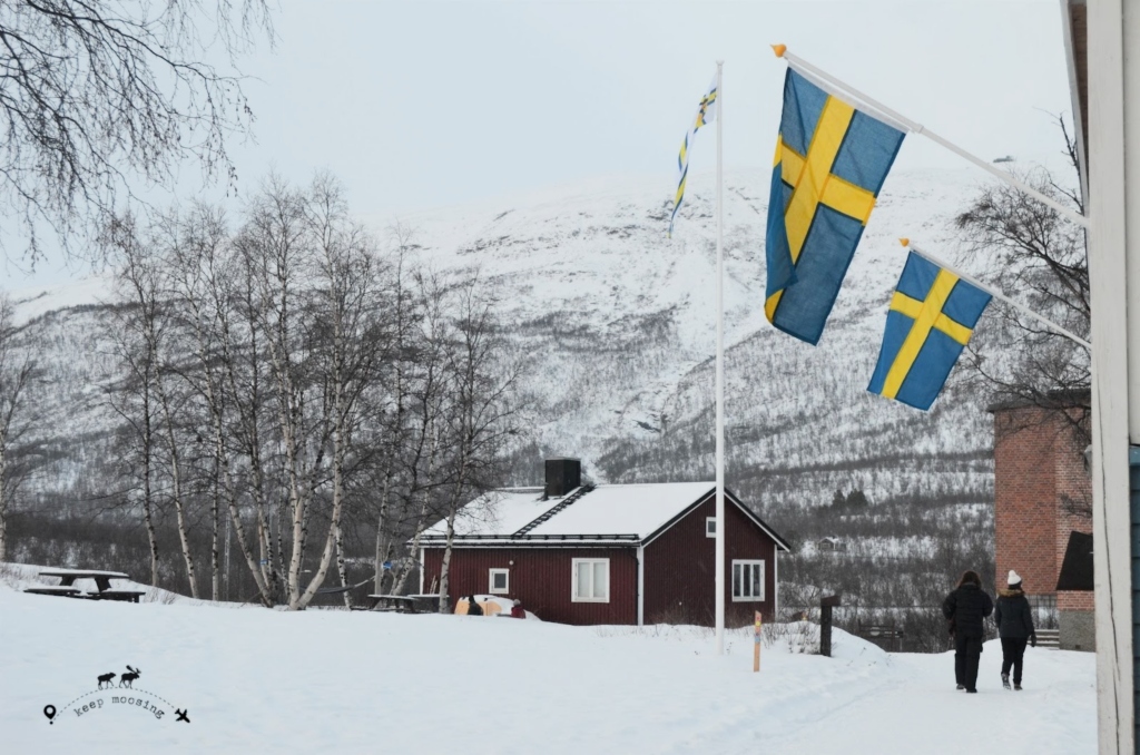 This article shows you what to see in Swedish Lapland traveling with us along our 5-day itinerary in this wonderful region. This image containing a red Swedish house in a snowy setting. Two Swedish flags are waving above two people walking in the snow.